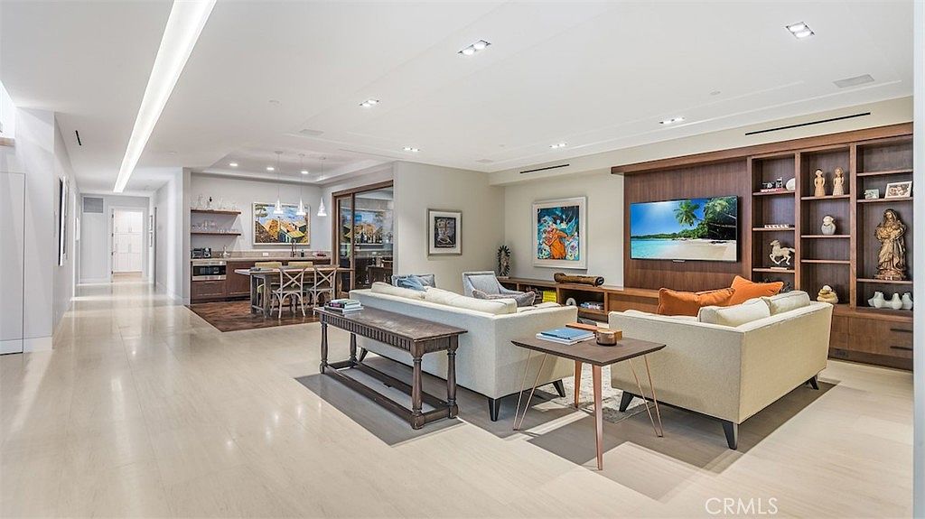 This spacious, open-concept living area features light-toned flooring and a neutral color palette, creating a bright and airy atmosphere. The room is anchored by a large, custom wood-paneled media wall with integrated shelving and a mounted television, while a comfortable seating arrangement faces the display. In the background, a dining area with a wooden table and chairs is visible, illuminated by modern pendant lights, providing a seamless transition between living and dining spaces.