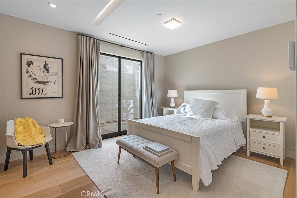 This bright and airy bedroom features a neutral color palette with light wood flooring and a large area rug. The room is furnished with a white bed frame, a matching nightstand with a lamp, and a small accent chair with a yellow throw blanket. A large glass sliding door provides access to an outdoor space, allowing natural light to fill the room.