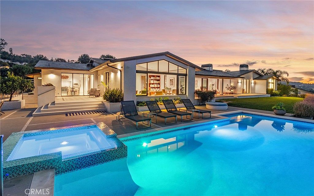This stunning rear exterior view showcases a modern single-story residence with a sprawling backyard featuring a large, illuminated swimming pool and a built-in spa. The home is characterized by its expansive glass walls that provide a seamless transition between the indoor living areas and the outdoor patio, which is furnished with several lounge chairs. The scene is captured at dusk, with a vibrant sunset sky casting a warm glow over the manicured lawn and the contemporary architectural lines of the house.