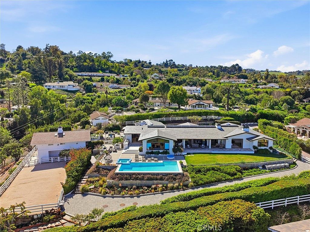 This high-angle aerial view captures a sprawling, single-story residence nestled within a lush, hilly landscape. The property features a prominent swimming pool on a stone-paved terrace, a detached garage structure, and expansive green lawns that blend into the surrounding natural terrain. The home's architecture is characterized by a low-profile design with multiple roof planes, creating a serene and private estate atmosphere.