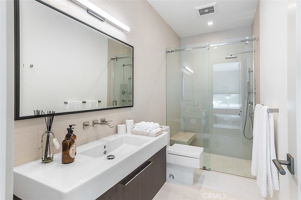This modern, minimalist bathroom features a floating vanity with a dark wood finish and a white countertop sink. A large, rectangular mirror hangs above the vanity, complemented by a sleek light fixture, while a glass-enclosed walk-in shower with a rainfall showerhead occupies the corner. The space is finished with light-toned wall tiles and a clean, contemporary aesthetic that creates a bright and airy atmosphere.