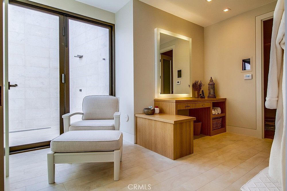 This serene primary bathroom features a light wood vanity with an integrated mirror and soft ambient lighting, creating a spa-like atmosphere. A comfortable lounge chair with an ottoman is positioned near large glass doors that open to a private outdoor space, allowing for natural light to fill the room. The space is finished with neutral-toned flooring and clean, minimalist lines, emphasizing a modern and tranquil aesthetic.