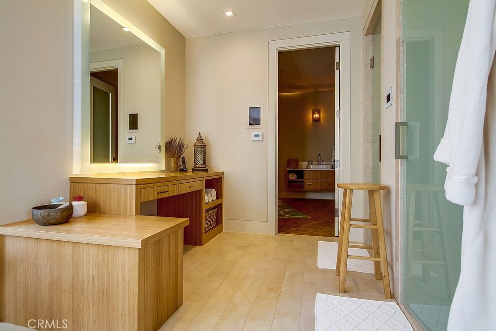 This bright and modern bathroom features a light wood vanity with a large, illuminated mirror and a decorative lantern accent. A wooden stool sits near a glass-enclosed shower, while a doorway leads into an adjacent space with a secondary vanity. The room is finished with light-toned flooring, creating a clean, spa-like atmosphere.