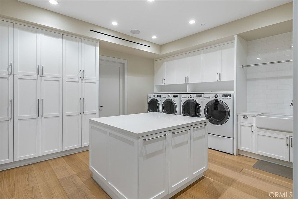 This spacious, modern laundry room features crisp white cabinetry, a central island with a white countertop, and light wood flooring. The room is equipped with two side-by-side washer and dryer sets, a farmhouse-style sink, and ample overhead storage. The bright, clean aesthetic is enhanced by recessed lighting and a neutral color palette, creating a highly functional and organized workspace.
