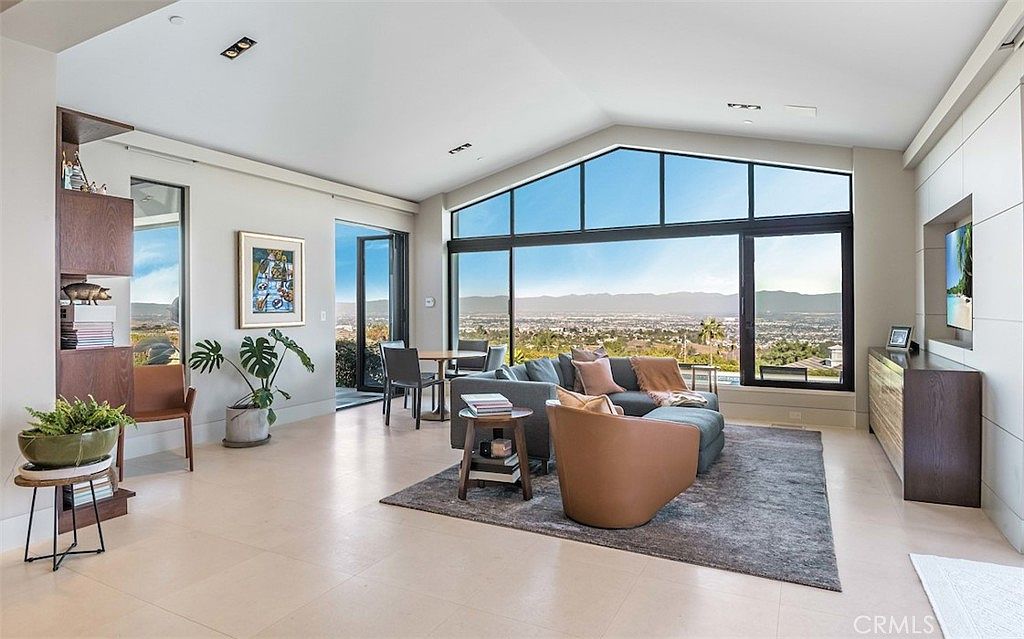 This spacious, contemporary living room features a large, angular window wall that frames an expansive panoramic view of the valley below. The room is furnished with a comfortable grey sectional sofa, a curved accent chair, and a minimalist wood-topped side table, all anchored by a dark area rug on light-toned tile flooring. A tall, built-in shelving unit on the left provides display space, while a sleek media console sits against the right wall, creating a sophisticated and airy atmosphere.