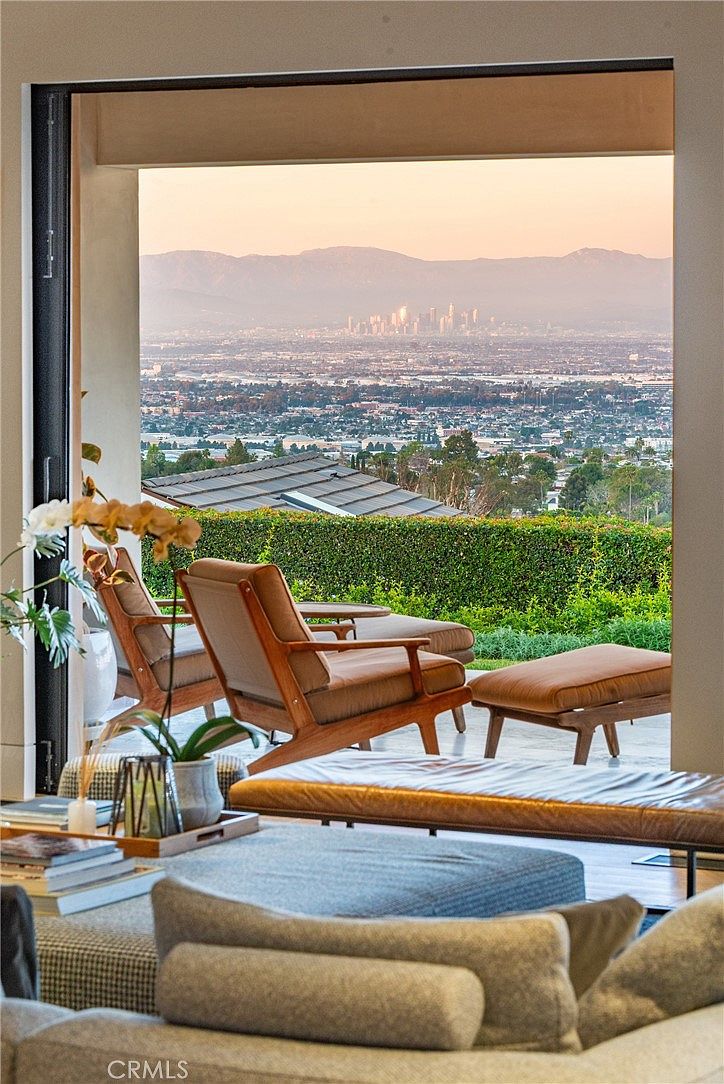 This interior living space features a seamless transition to an outdoor patio through expansive, floor-to-ceiling glass doors that frame a panoramic city and mountain view. The foreground includes a comfortable seating area with a leather bench and plush sofa, while the patio is furnished with two wooden lounge chairs and an ottoman. The overall aesthetic is sophisticated and serene, emphasizing the connection between the indoor living area and the scenic outdoor landscape.