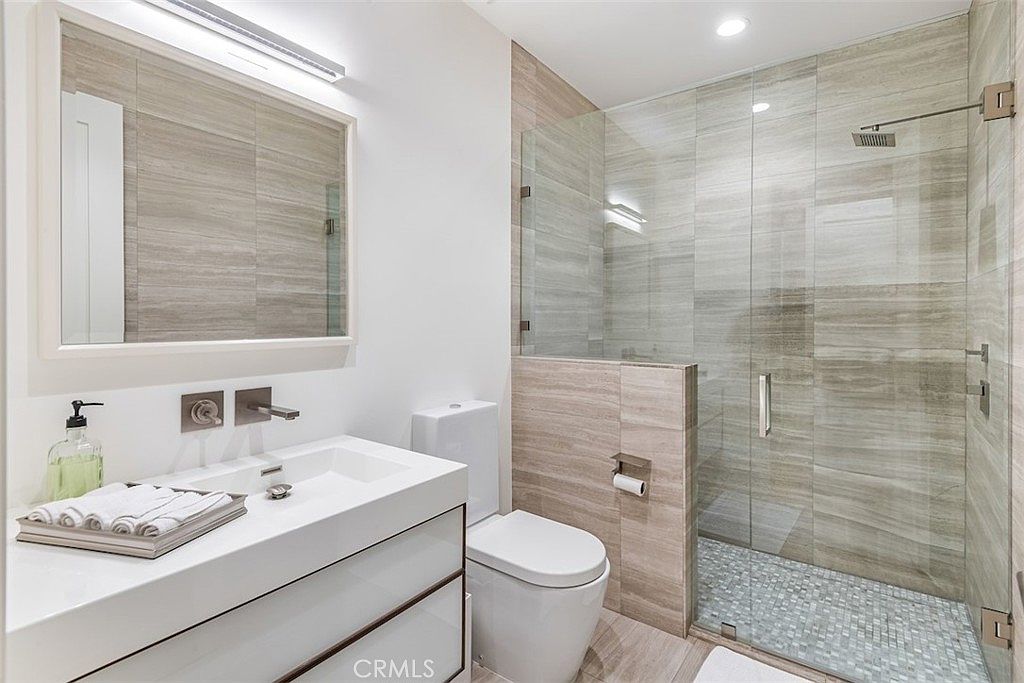 This modern bathroom features a sleek vanity with a rectangular sink and a large, framed mirror above it. To the right, a walk-in shower is enclosed by glass panels and finished with light-toned, horizontal-patterned wall tiles and a mosaic-tiled floor. The space is bright and minimalist, utilizing a neutral color palette to create a clean, contemporary aesthetic.