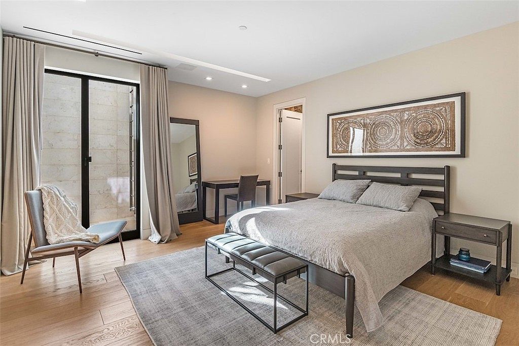 This contemporary bedroom features light wood flooring, neutral wall tones, and a large bed centered against a wall adorned with a framed piece of textured art. To the left, a floor-to-ceiling glass door provides access to an outdoor space, while a full-length mirror and a small desk area are positioned nearby. The room is illuminated by recessed ceiling lights, creating a clean, minimalist, and inviting atmosphere.