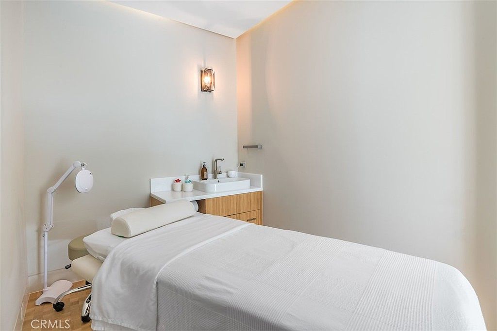 This image depicts a professional spa or treatment room featuring a massage table covered in white linens and a bolster pillow. In the background, a built-in vanity with a sink and light wood cabinetry provides a functional workspace, complemented by a wall-mounted light fixture. The room has a neutral color palette, creating a calm and serene atmosphere suitable for wellness services.
