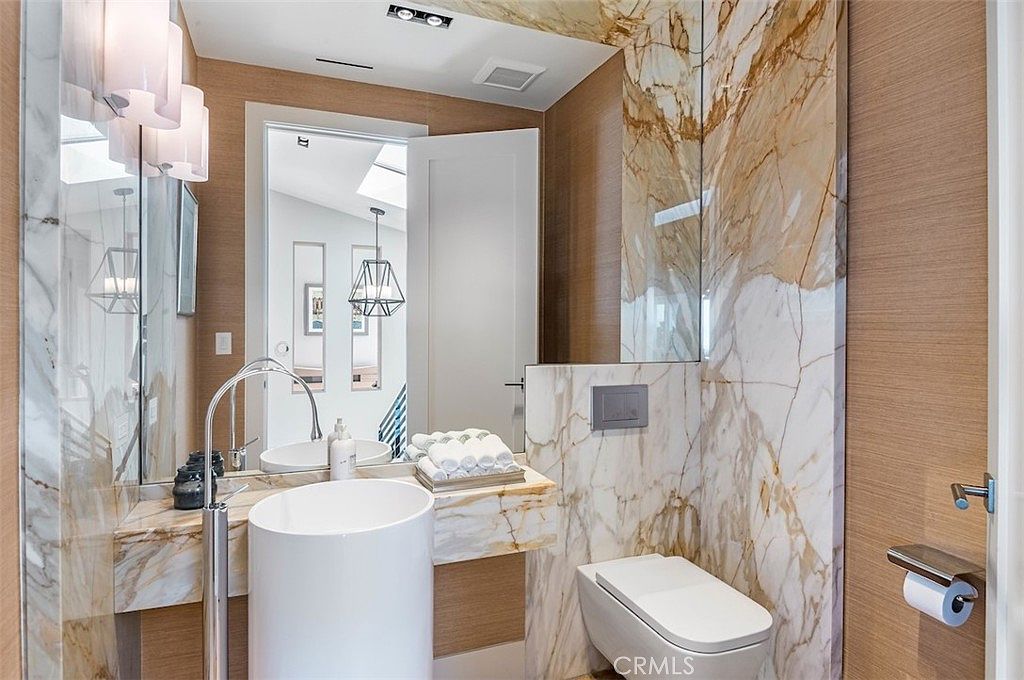 This modern powder room features a striking wall of veined stone that extends behind the wall-mounted toilet and vanity area. A cylindrical pedestal sink with a tall, curved faucet stands as a focal point, complemented by a large mirror and warm-toned wood paneling on the side walls. The space is illuminated by sleek, vertical light fixtures, creating a sophisticated and high-end aesthetic.