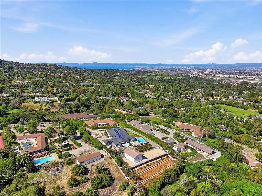 This high-angle aerial view captures a sprawling residential neighborhood nestled in a lush, green landscape with a distant view of the ocean. The property features a large, modern-style home with a dark roof, a swimming pool, and extensive grounds including a fenced area and mature trees. The perspective provides a comprehensive look at the property's layout and its integration into the surrounding suburban environment.