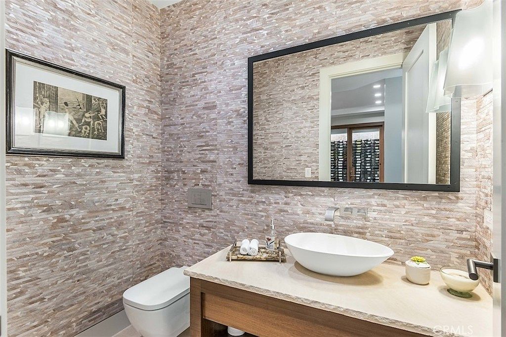 This modern powder room features a striking stacked-stone wall texture that provides a sophisticated, organic backdrop. A vessel sink sits atop a light-toned stone vanity, complemented by a large, dark-framed mirror and a wall-mounted faucet. The space is finished with a sleek, wall-mounted toilet and warm, ambient lighting, creating an elegant and contemporary atmosphere.