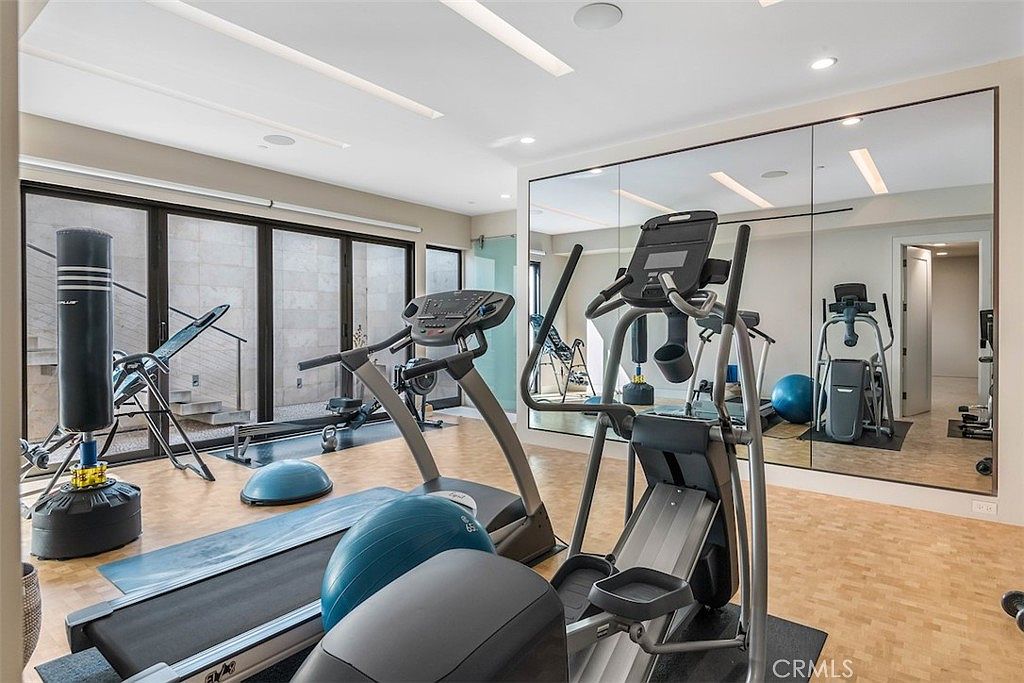 This spacious home gym features a variety of fitness equipment, including a treadmill, elliptical, and a punching bag. The room is accented by a large wall mirror that enhances the sense of space, while light wood flooring provides a clean, functional surface. Large glass doors lead to an outdoor area, allowing natural light to fill the room and creating an inviting atmosphere for exercise.