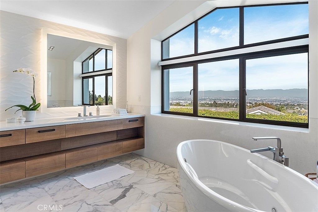 This primary bathroom features a sleek, floating vanity with light wood cabinetry and a large, illuminated mirror. A freestanding soaking tub is positioned near a expansive window that offers a panoramic view of the surrounding landscape. The space is finished with light-toned, patterned stone flooring, creating a bright and luxurious atmosphere.