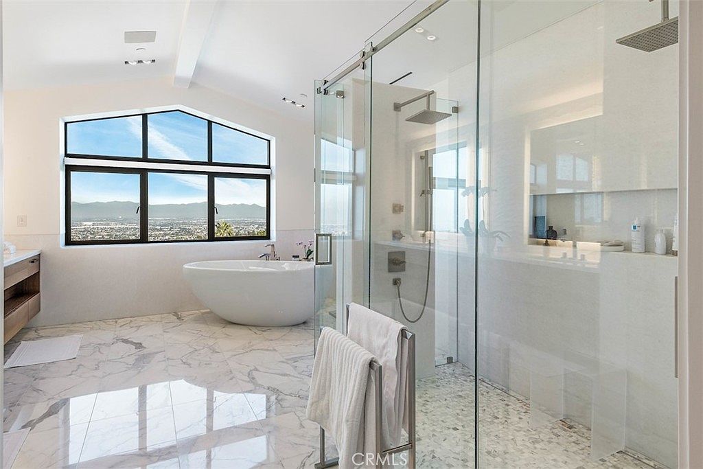 This luxurious primary bathroom features a freestanding soaking tub positioned beneath a large window that offers expansive views. A spacious walk-in shower with glass walls and a rainfall showerhead occupies the center of the room, complemented by light-toned stone flooring throughout. The space is bright and airy, emphasizing a clean, modern aesthetic with high-end finishes.