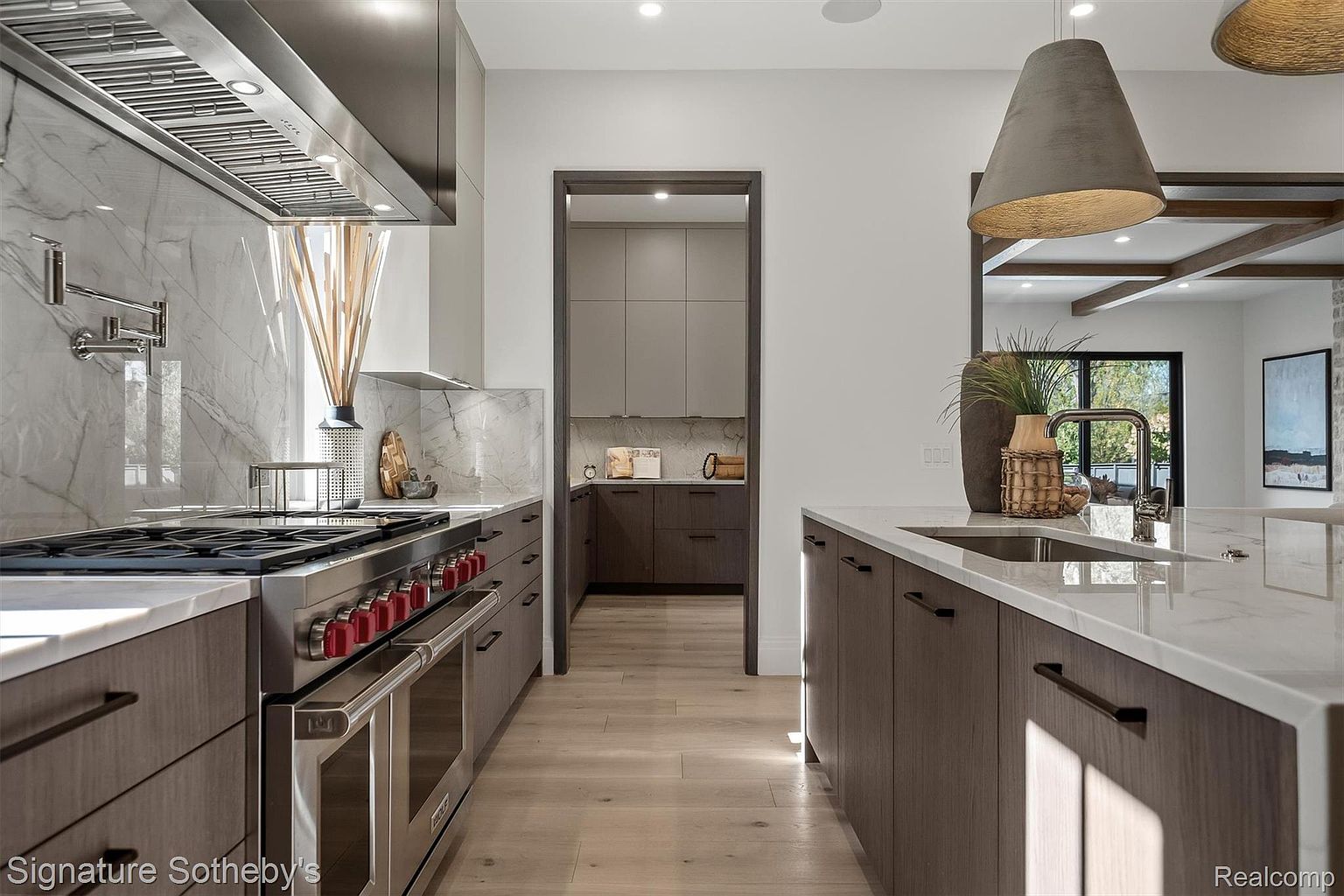 This is a modern kitchen with a focus on clean lines and high-end appliances. The kitchen features a large island with a sink, custom cabinetry with dark hardware, and a professional-grade range with a stainless steel hood. A doorway leads to a pantry area, and the overall impression is one of luxury and functionality.