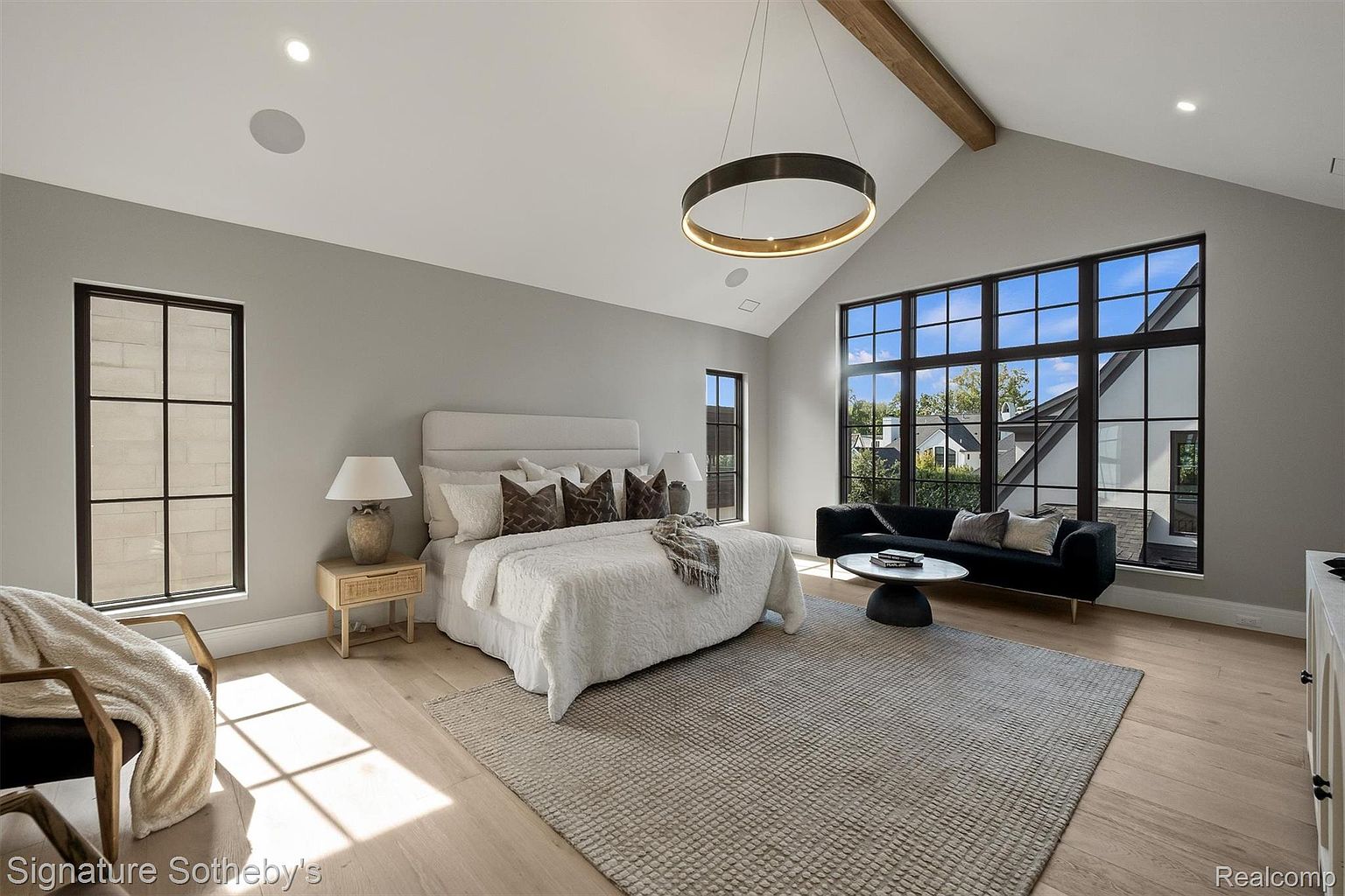 This is a bright and spacious primary bedroom featuring a large bed with a white comforter and decorative pillows, complemented by a cozy seating area with a dark sofa and round coffee table. The room is well-lit by natural light streaming through large windows with black frames, and a modern circular chandelier hangs from the vaulted ceiling with a wooden beam. The overall aesthetic is modern and luxurious, creating a serene and inviting atmosphere.