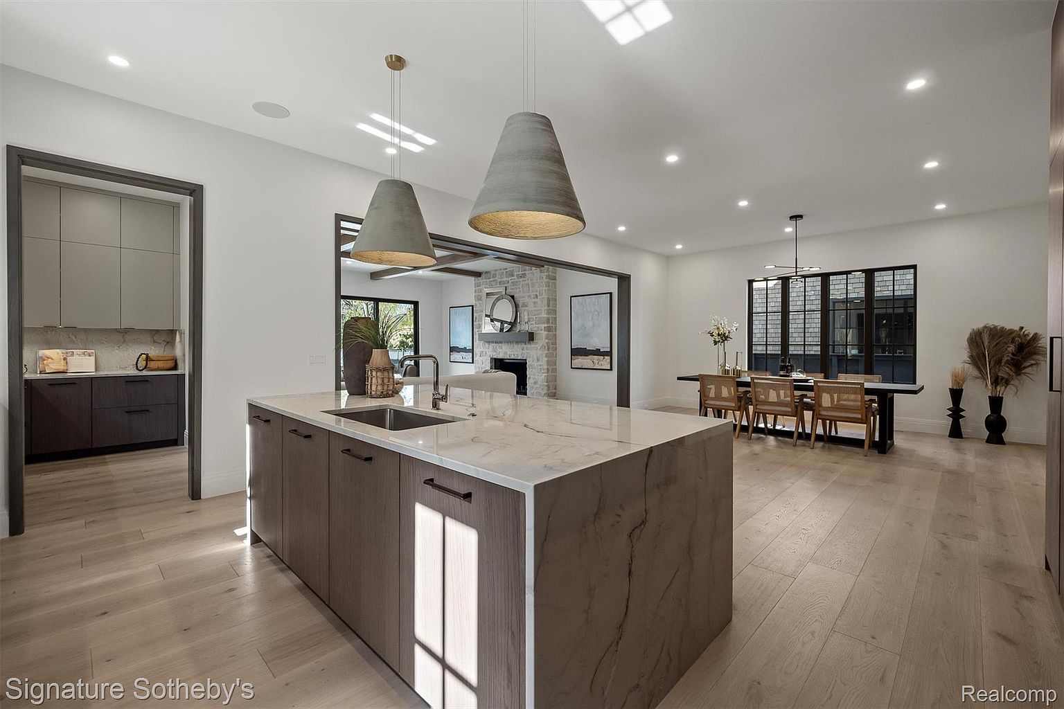 This is a bright and modern kitchen featuring a large island with a marble countertop and dark wood cabinetry. Two pendant lights hang above the island, and the open floor plan connects the kitchen to the dining area and living room with a fireplace. The hardwood floors and neutral color palette create a warm and inviting atmosphere.