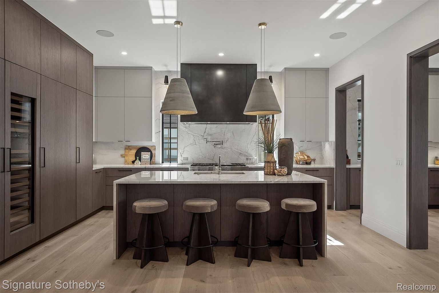 This is a modern kitchen featuring a large island with seating for four, dark wood cabinetry, and light gray upper cabinets. The kitchen also has marble countertops and backsplash, stainless steel appliances, and pendant lighting. The overall impression is one of luxury and sophistication.