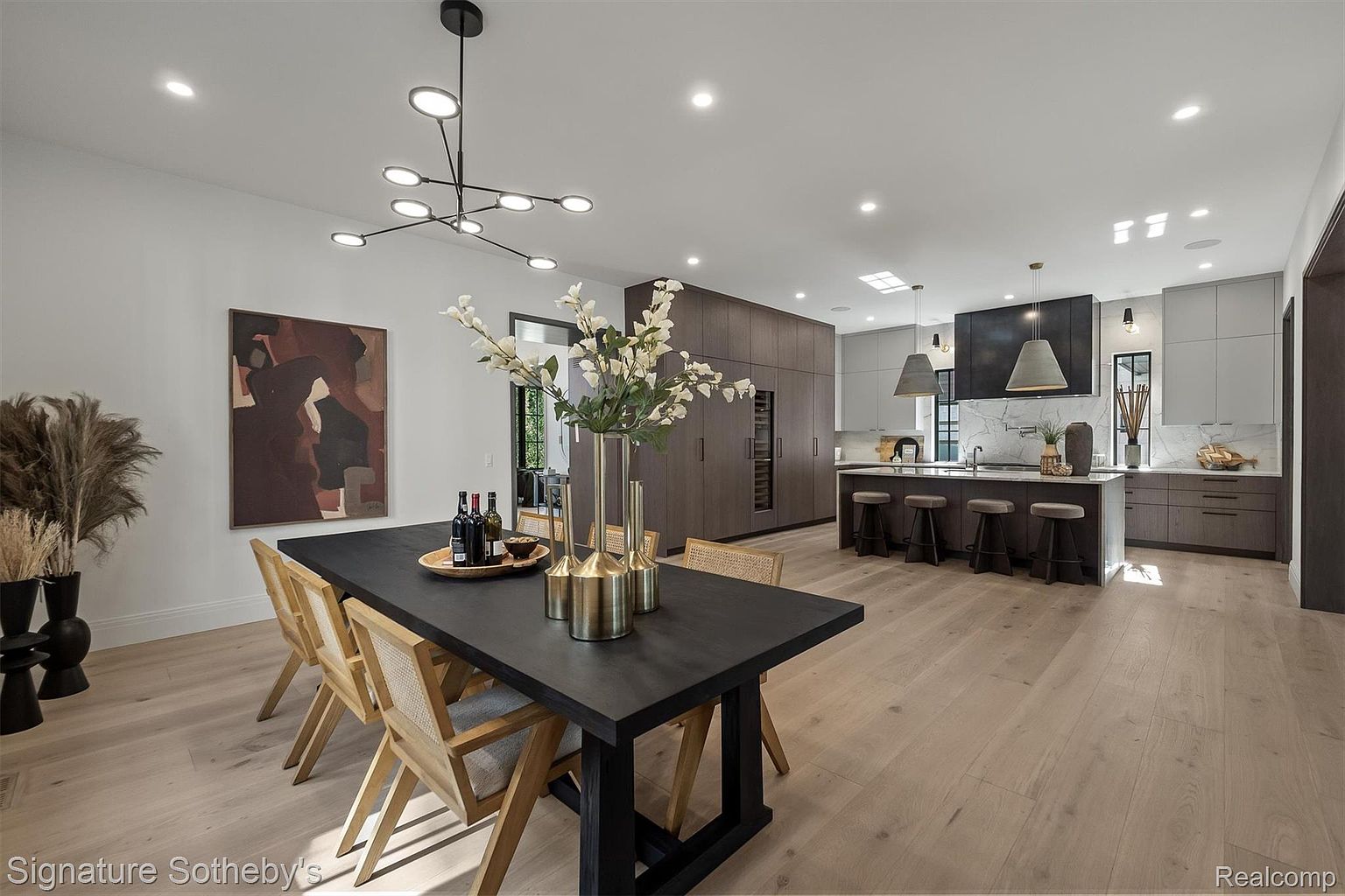 This interior shot showcases a modern dining room that seamlessly transitions into a contemporary kitchen. The dining area features a dark wood table surrounded by light wood chairs, complemented by a striking chandelier and abstract art. The kitchen boasts sleek cabinetry, a central island with bar stools, and modern pendant lighting, creating an open and inviting space perfect for entertaining.