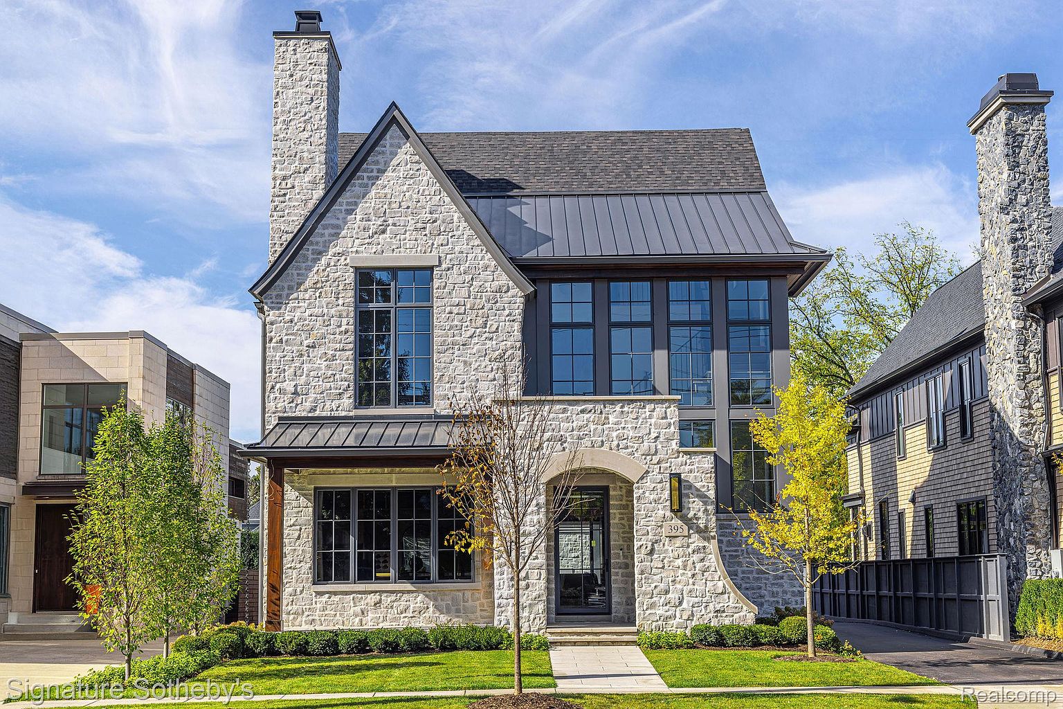 This is a front exterior view of a two-story stone house with a metal roof and black-framed windows. The house features a prominent chimney, a well-manicured lawn, and mature trees. The overall impression is one of sophisticated elegance and curb appeal, making it a desirable property.