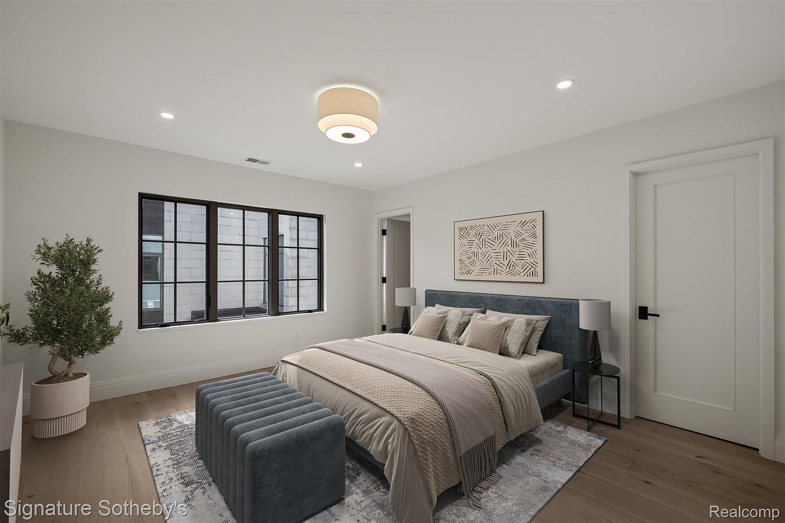 This is a well-lit primary bedroom featuring a large window with black frames, a bed with a blue headboard, and a neutral-toned comforter. An ottoman sits at the foot of the bed, and a modern art piece hangs on the wall. The room has a clean, contemporary aesthetic with hardwood floors and white walls.