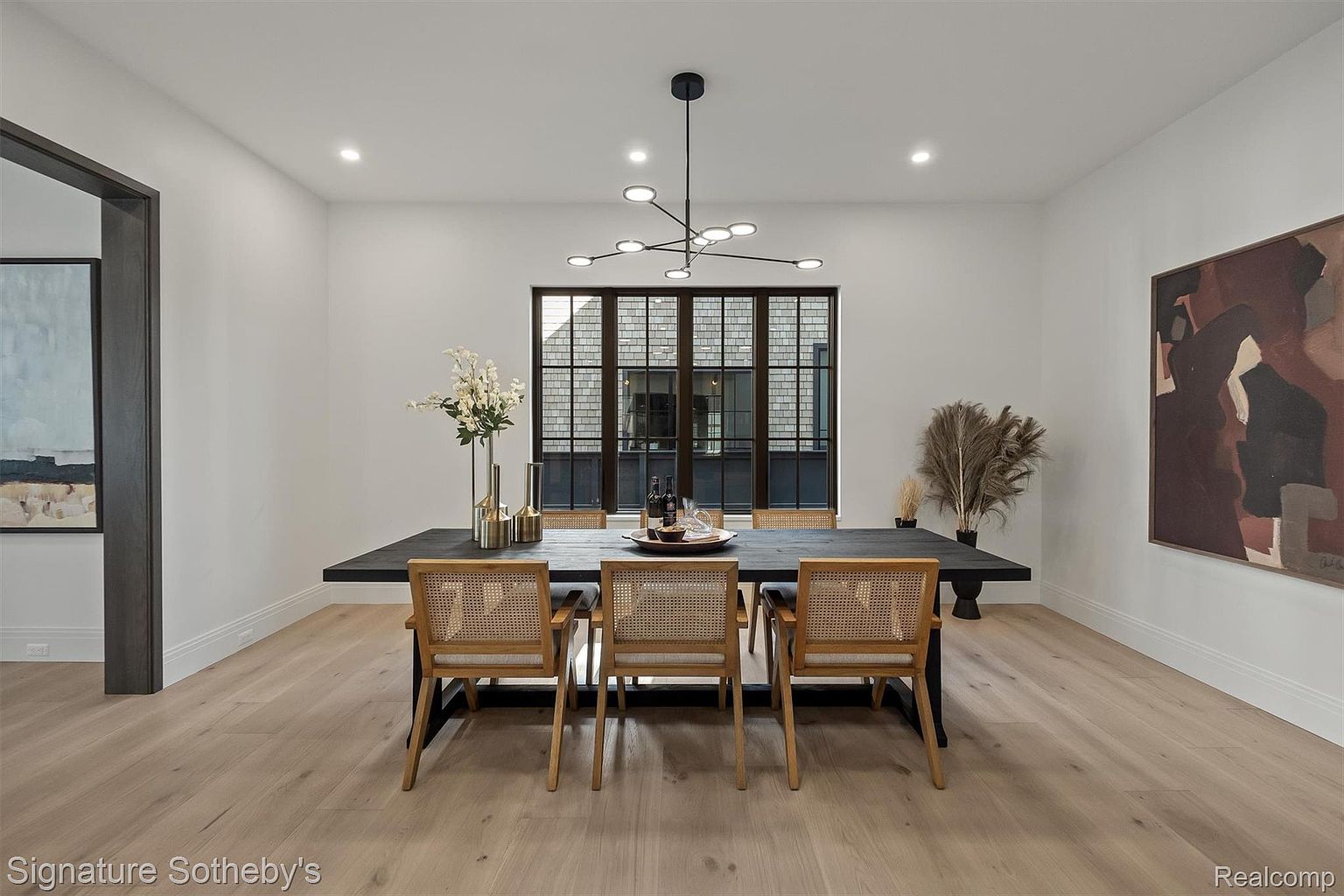 This is a dining room featuring a long, dark wood table with seating for six. The chairs have woven backs and light wood frames. A modern chandelier hangs above the table, and a large window provides natural light. Artwork adorns the walls, and the overall style is contemporary and elegant.