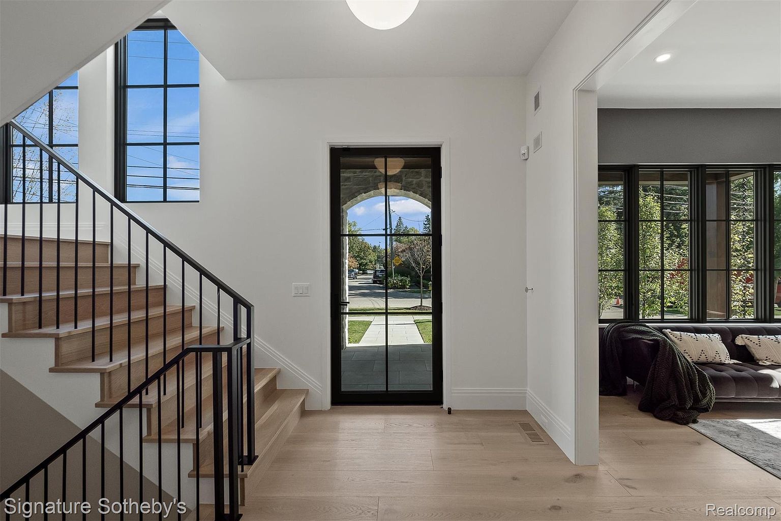 This interior shot showcases a bright and modern entryway with a staircase. The light wood flooring complements the white walls, while black-framed windows and a front door add a contemporary touch. The staircase features wooden steps and a black metal railing, leading to the upper level.