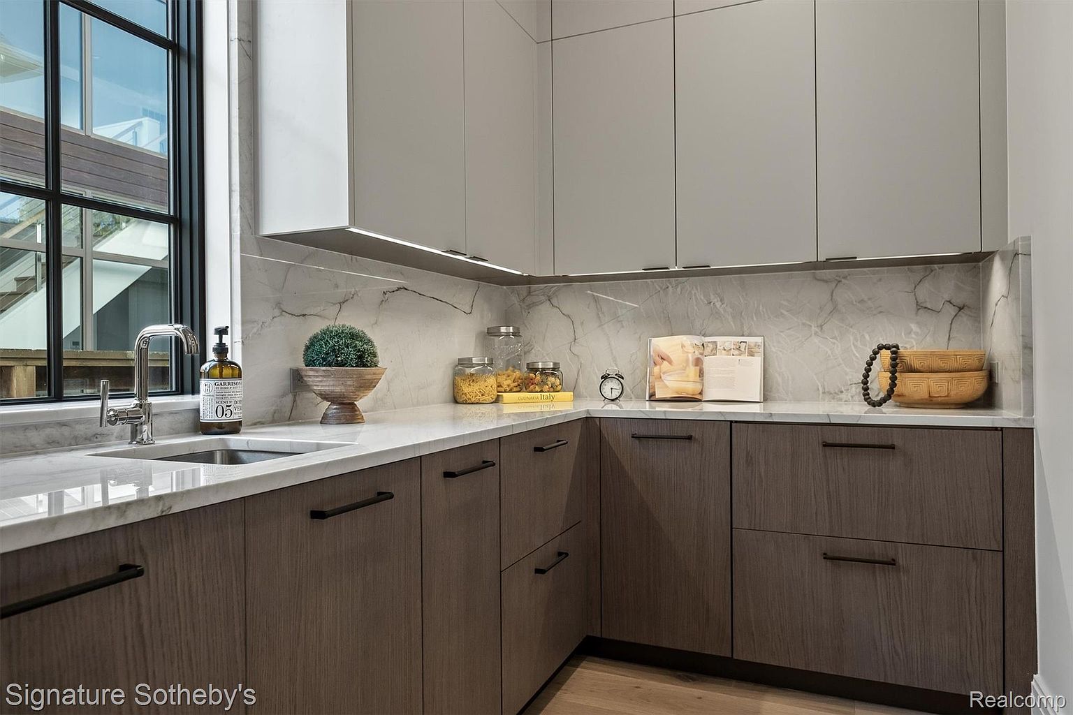 This is a well-lit kitchen featuring two-tone cabinetry with light gray upper cabinets and wood-grain lower cabinets. The countertops and backsplash are a light marble, and a stainless steel sink is visible near a window. The kitchen is decorated with various items, including jars, bowls, and a cookbook, creating a warm and inviting atmosphere.