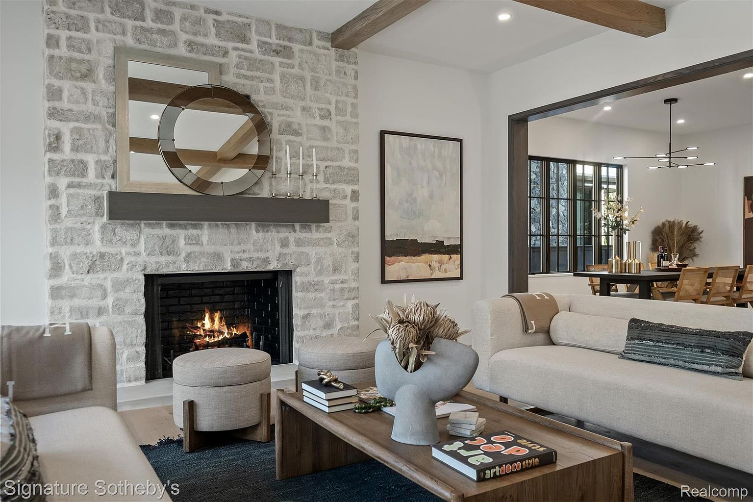 This interior shot showcases a cozy living room with a stone fireplace as the focal point, complemented by a wooden mantel and decorative mirror. The room features neutral-toned furniture, including a sofa and ottomans, arranged around a wooden coffee table adorned with books and a unique vase. The open layout leads to a dining area, creating a sense of spaciousness and modern comfort.