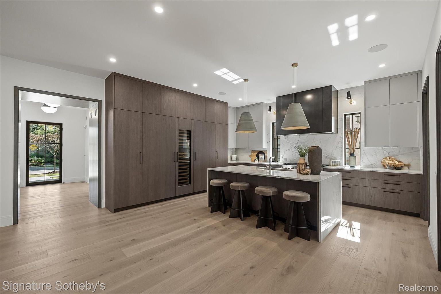 This is a modern kitchen with light wood flooring, a large island with seating, and a combination of dark wood and light gray cabinetry. The kitchen features a wine fridge, marble backsplash, and pendant lighting, creating a luxurious and functional space. The open layout and natural light enhance the overall appeal.