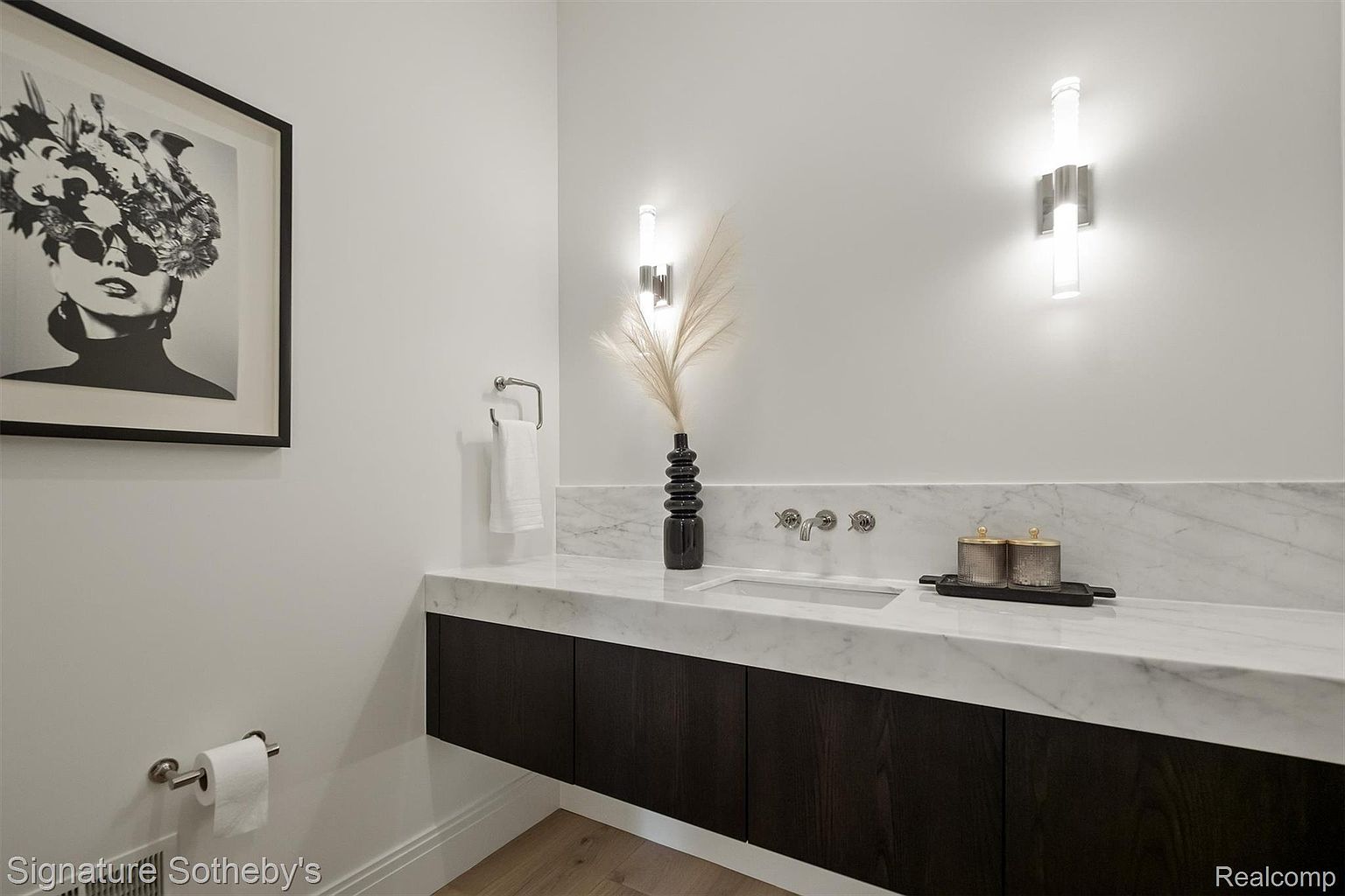 This is a modern guest bathroom featuring a floating dark wood vanity with a white marble countertop and backsplash. The walls are painted a clean white, and the room is illuminated by contemporary sconces. A framed black and white art piece adds a touch of sophistication to the space.