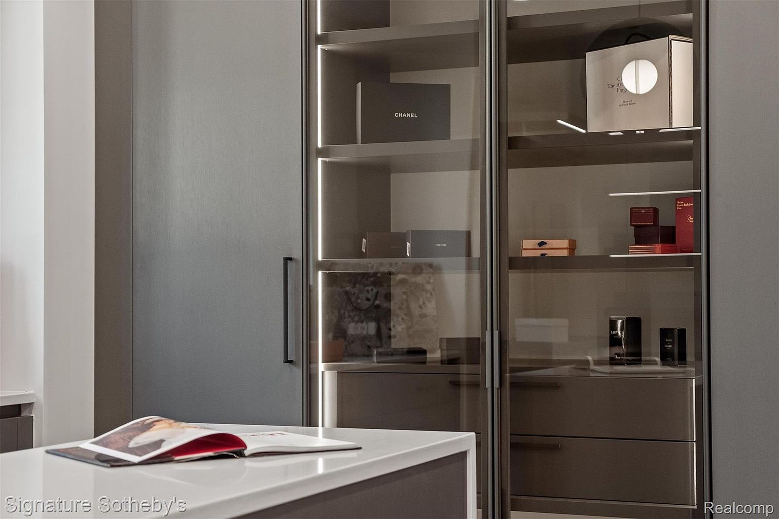 This image showcases a luxurious walk-in closet with sleek, modern design elements. The closet features glass-fronted cabinets with integrated lighting, displaying designer boxes and accessories. A central island with a light countertop provides a space for organizing and displaying items, enhancing the closet's functionality and aesthetic appeal.
