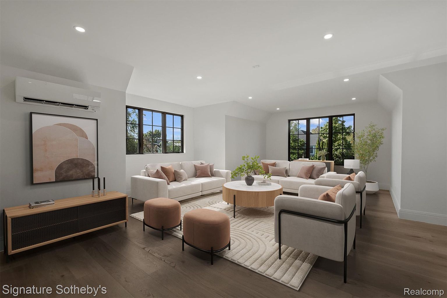 This is a bright and airy living room featuring two white sofas with decorative pillows, two armchairs, and two brown ottomans arranged around a round coffee table on a patterned rug. Large windows provide natural light, and the hardwood floors add warmth to the space. A modern console table with artwork sits against one wall, enhancing the room's contemporary style.