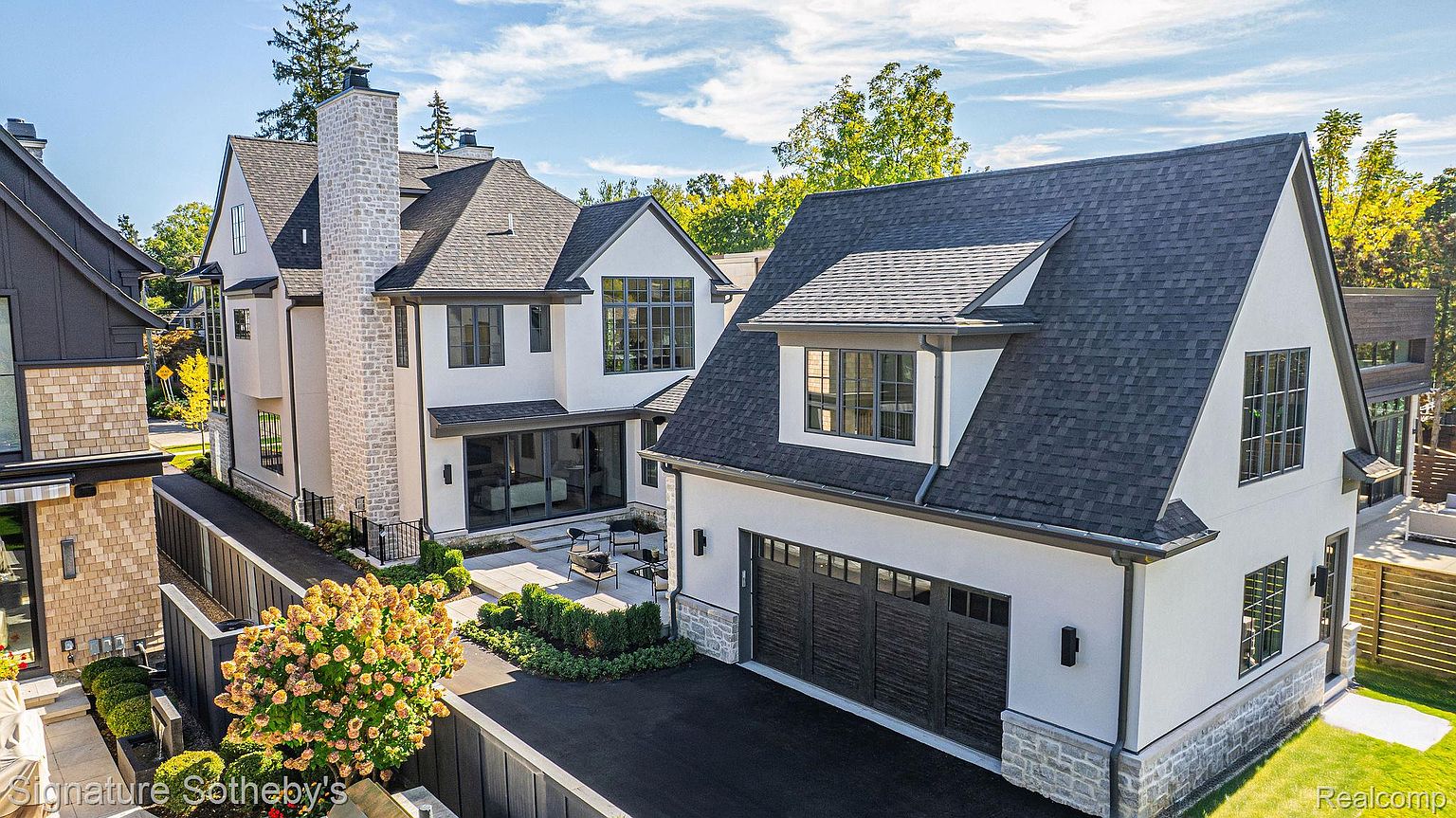 This aerial shot showcases a luxurious modern home with a sophisticated exterior design. The property features a combination of light stucco and stone accents, complemented by dark-framed windows and a dark shingle roof. A well-manicured yard and paved driveway add to the overall curb appeal, highlighting the property's high-end aesthetic.