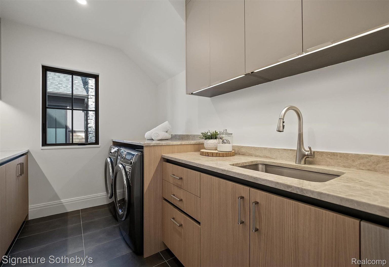 This is a well-appointed laundry room featuring modern cabinetry with light wood finishes and sleek hardware. A stainless steel sink is integrated into the countertop, complemented by a stylish faucet. The room also includes a front-loading washer and dryer set, and a window provides natural light, enhancing the clean and functional design.