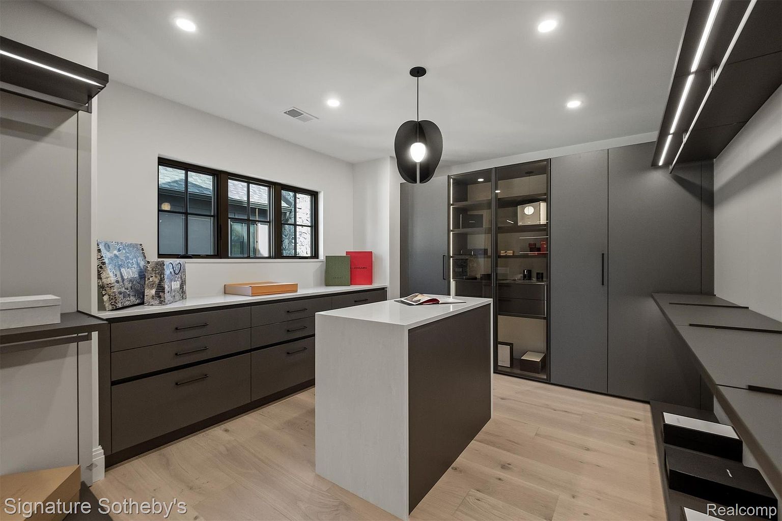 This is a well-organized walk-in closet featuring custom cabinetry in a dark gray finish with contrasting white countertops and island. The closet includes drawers, shelving, and glass-fronted display cabinets, illuminated by modern lighting fixtures. The overall impression is luxurious and functional, designed for efficient storage and display of clothing and accessories.