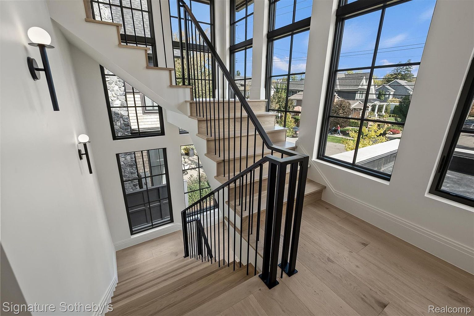 This interior shot showcases a modern staircase with light wood treads and black metal railings, ascending alongside a wall adorned with sconce lighting and large windows. The windows offer a view of the neighborhood outside, and the light wood flooring complements the overall contemporary design. The perspective is from above, looking down the staircase.