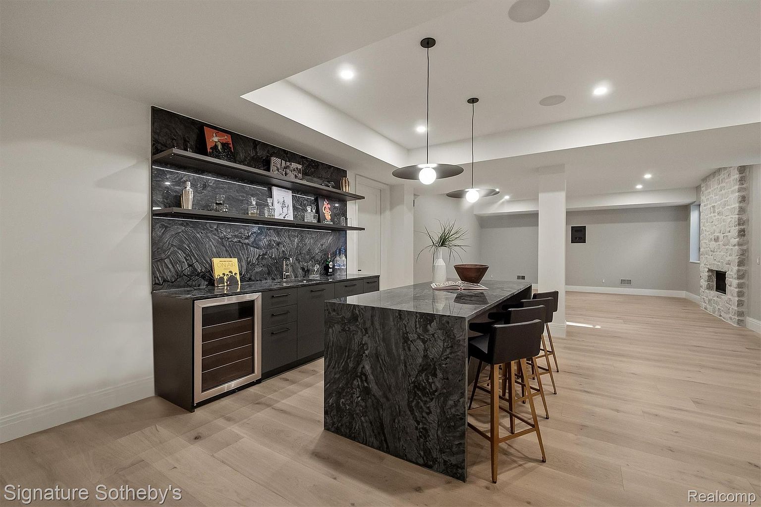 This interior shot showcases a stylish basement area featuring a wet bar with dark cabinetry, marble backsplash, and open shelving. A matching island with bar seating anchors the space, complemented by modern pendant lighting. The room extends into a spacious area with light wood flooring and a stone fireplace, creating a sophisticated and inviting atmosphere.
