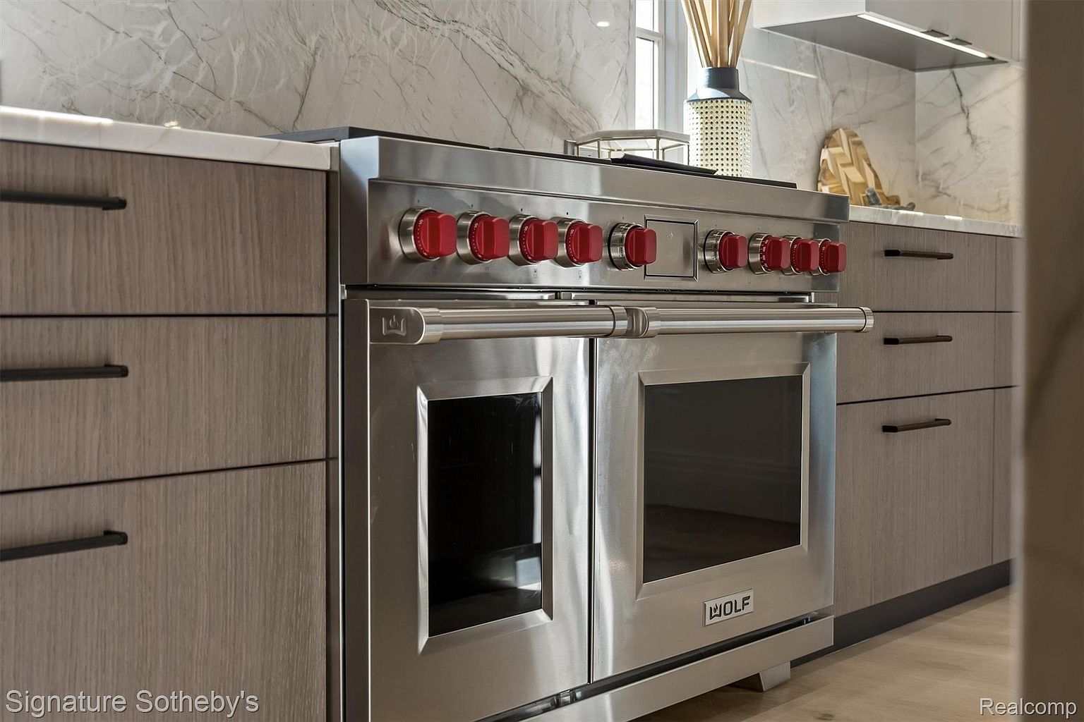 This is a detailed shot of a modern kitchen featuring a high-end stainless steel Wolf range with red knobs, set against a backdrop of sleek, wood-grain cabinetry with minimalist hardware and a marble backsplash. The focus is on the appliance, showcasing its professional-grade quality and design, which adds a luxurious touch to the kitchen space.
