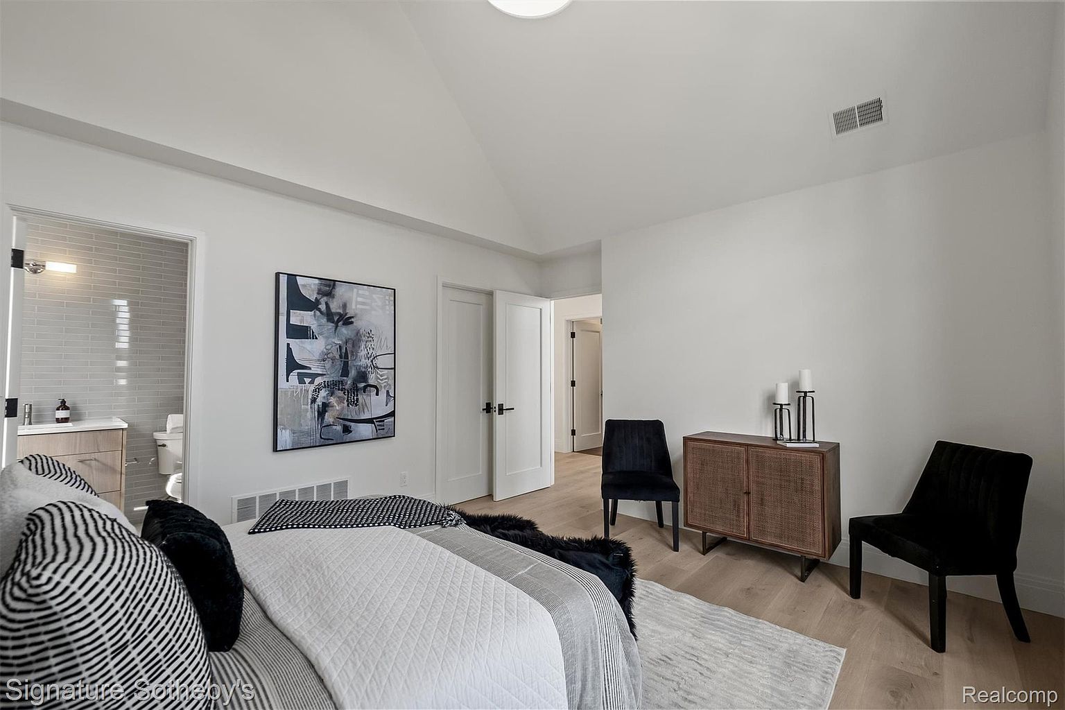This is a primary bedroom featuring a modern design with white walls and light wood flooring. The room includes a bed with stylish bedding, an abstract art piece, and two black chairs flanking a wooden cabinet with decorative candles. The space is well-lit and appears spacious, with doorways leading to other areas of the house.