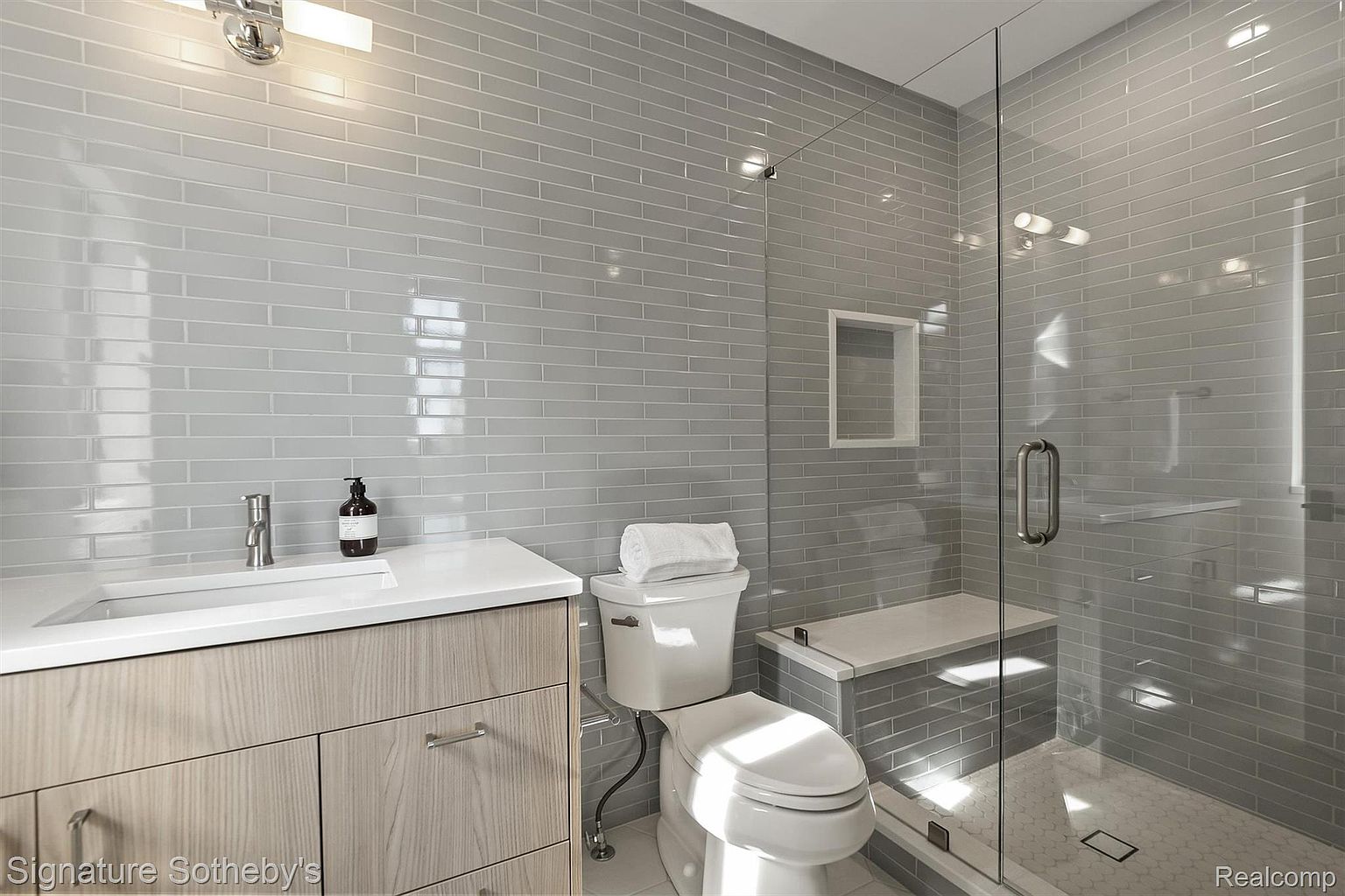 This is a primary bathroom featuring a modern design with gray subway tile walls and a glass-enclosed shower. The vanity has a light wood finish with a white countertop, and a toilet is positioned next to the shower. The shower includes a built-in bench and a niche for toiletries, enhancing the spa-like atmosphere.