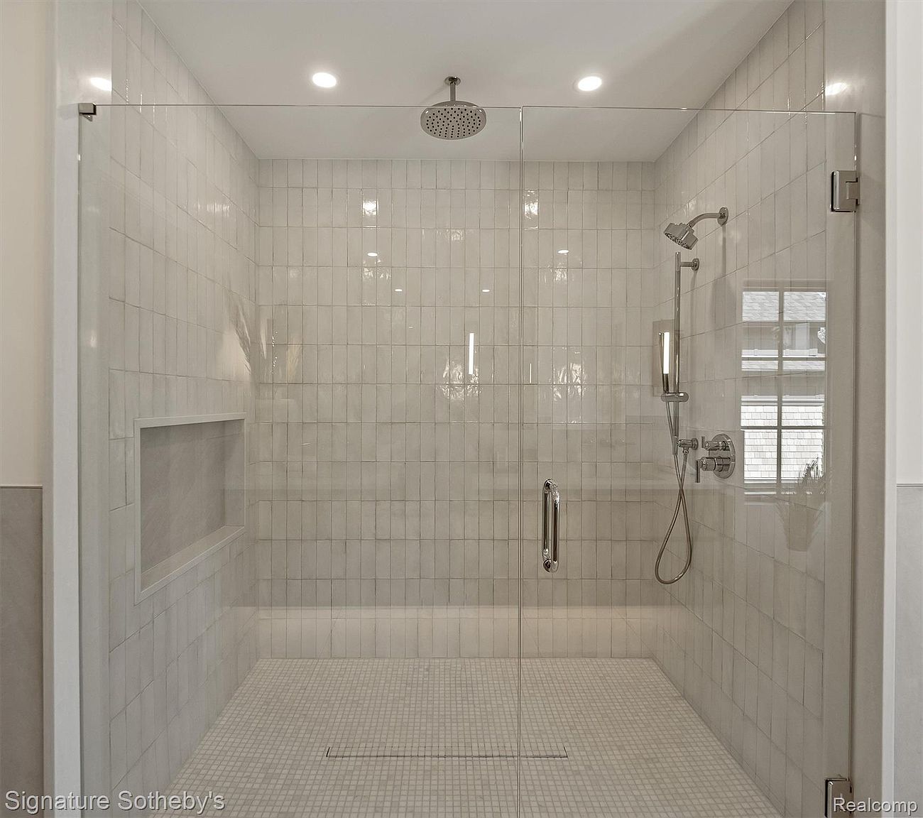 This is a well-lit primary bathroom featuring a modern, glass-enclosed shower. The shower is tiled with light-colored, vertically stacked rectangular tiles and has a mosaic tile floor. The shower includes both a rainfall showerhead and a handheld shower fixture, along with a built-in niche for storage.