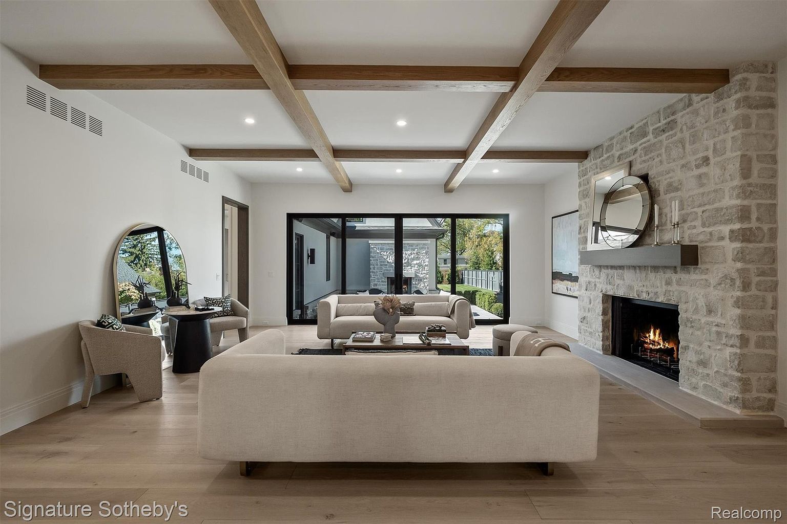 This is a bright and spacious living room featuring a modern design with exposed wooden beams on the ceiling. The room is furnished with comfortable sofas and chairs, centered around a fireplace with a stone surround. Large sliding glass doors provide ample natural light and a view of the outdoor space, creating an inviting and luxurious atmosphere.