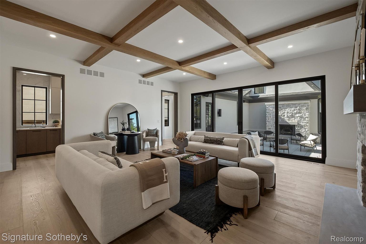 This is an interior shot of a modern living room featuring a neutral color palette with wooden beam accents on the ceiling. The room is furnished with two light-colored sofas, ottomans, a dark rug, and a wooden coffee table. Large sliding glass doors provide access to an outdoor patio area with a fireplace, creating a seamless indoor-outdoor living experience.
