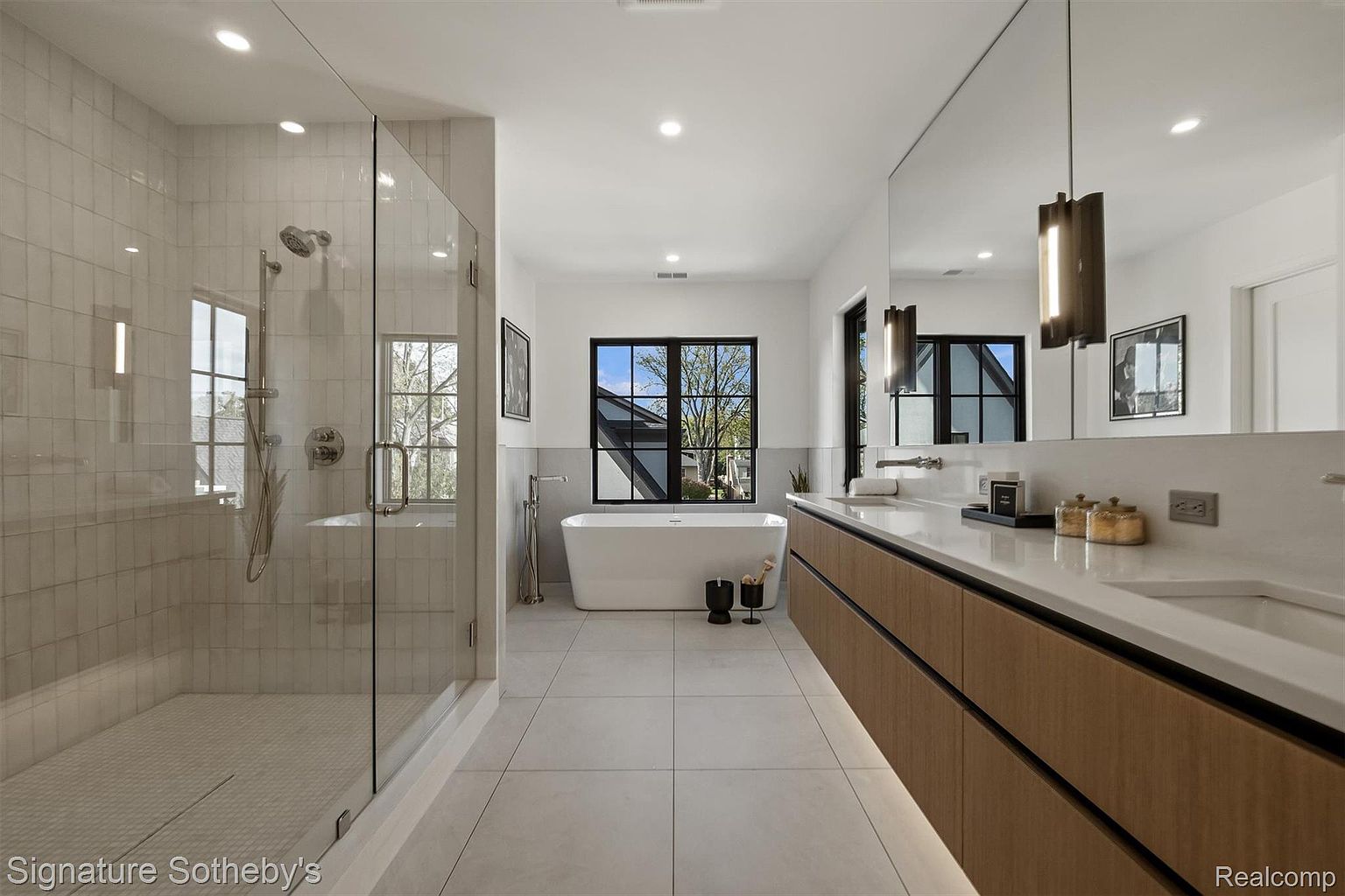 This is a luxurious primary bathroom featuring a glass-enclosed shower with white tile, a freestanding soaking tub under a large window, and a modern vanity with wood-grain cabinets and a white countertop. The room is well-lit with recessed lighting and pendant fixtures, creating a spa-like atmosphere. The overall impression is clean, contemporary, and upscale.