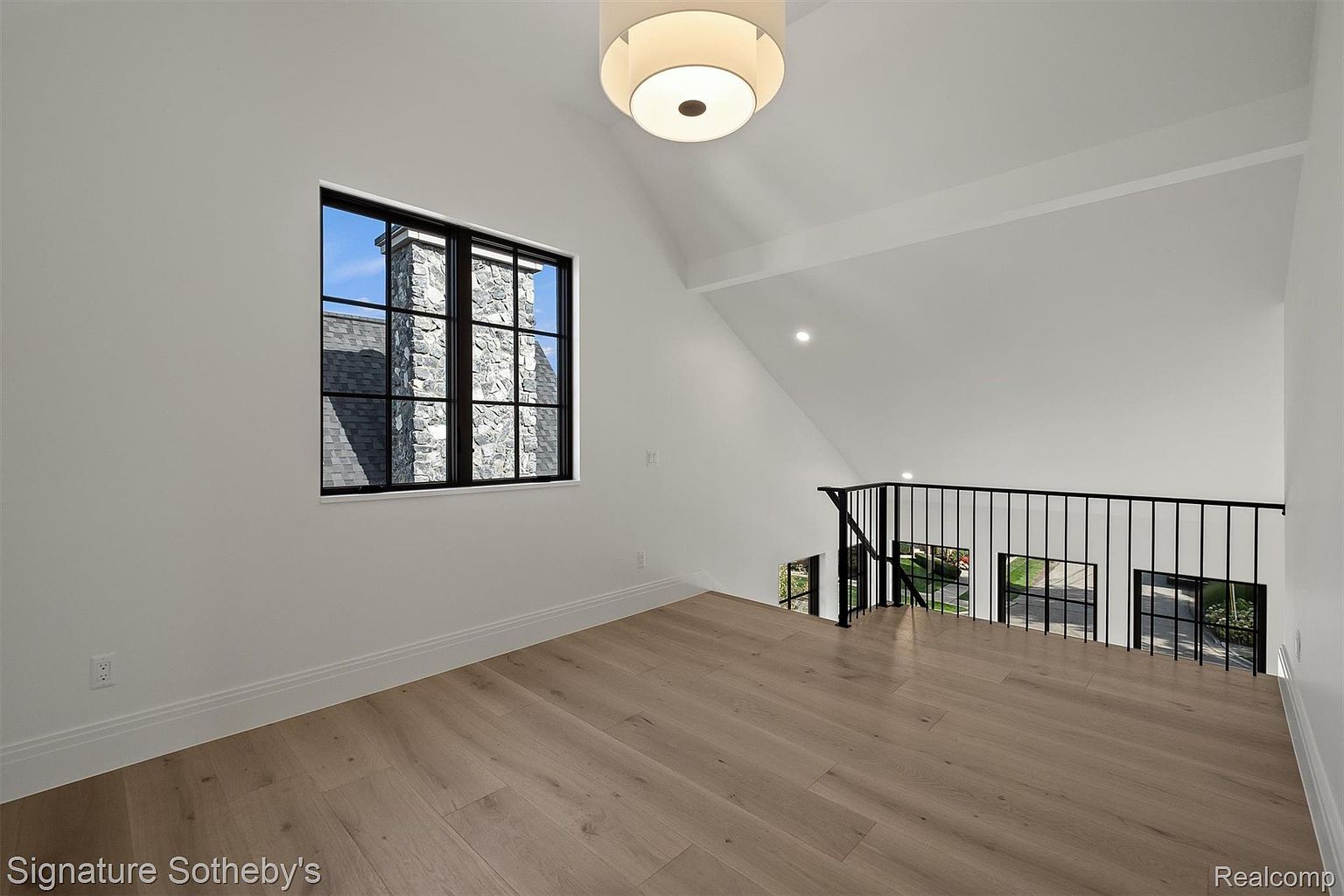 This interior shot showcases a bright hallway or landing area with light wood flooring and white walls, creating a clean and spacious feel. A black metal railing overlooks a lower level, and a window provides natural light and a view of the exterior. A modern ceiling light fixture illuminates the space, enhancing the contemporary design.