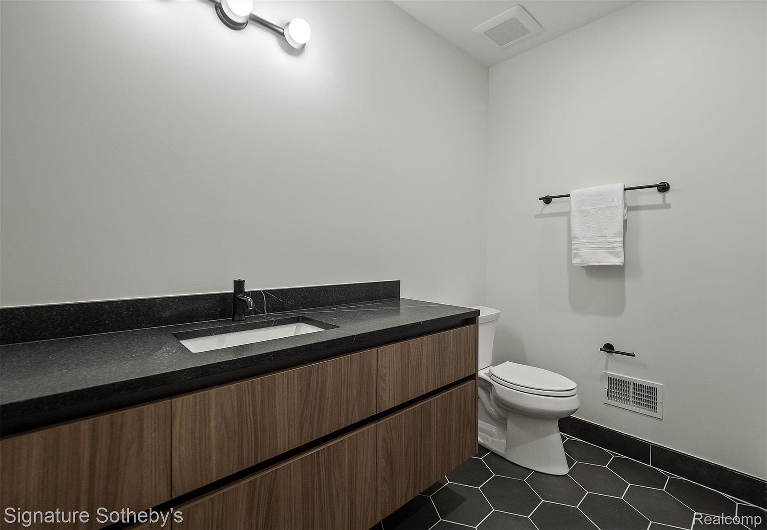 This is a modern bathroom featuring a dark countertop with a sleek, integrated sink and a wood-grain vanity. The black hexagonal floor tiles add a contemporary touch, while the white walls and minimalist fixtures create a clean and sophisticated look. A white towel hangs on a black towel rack.
