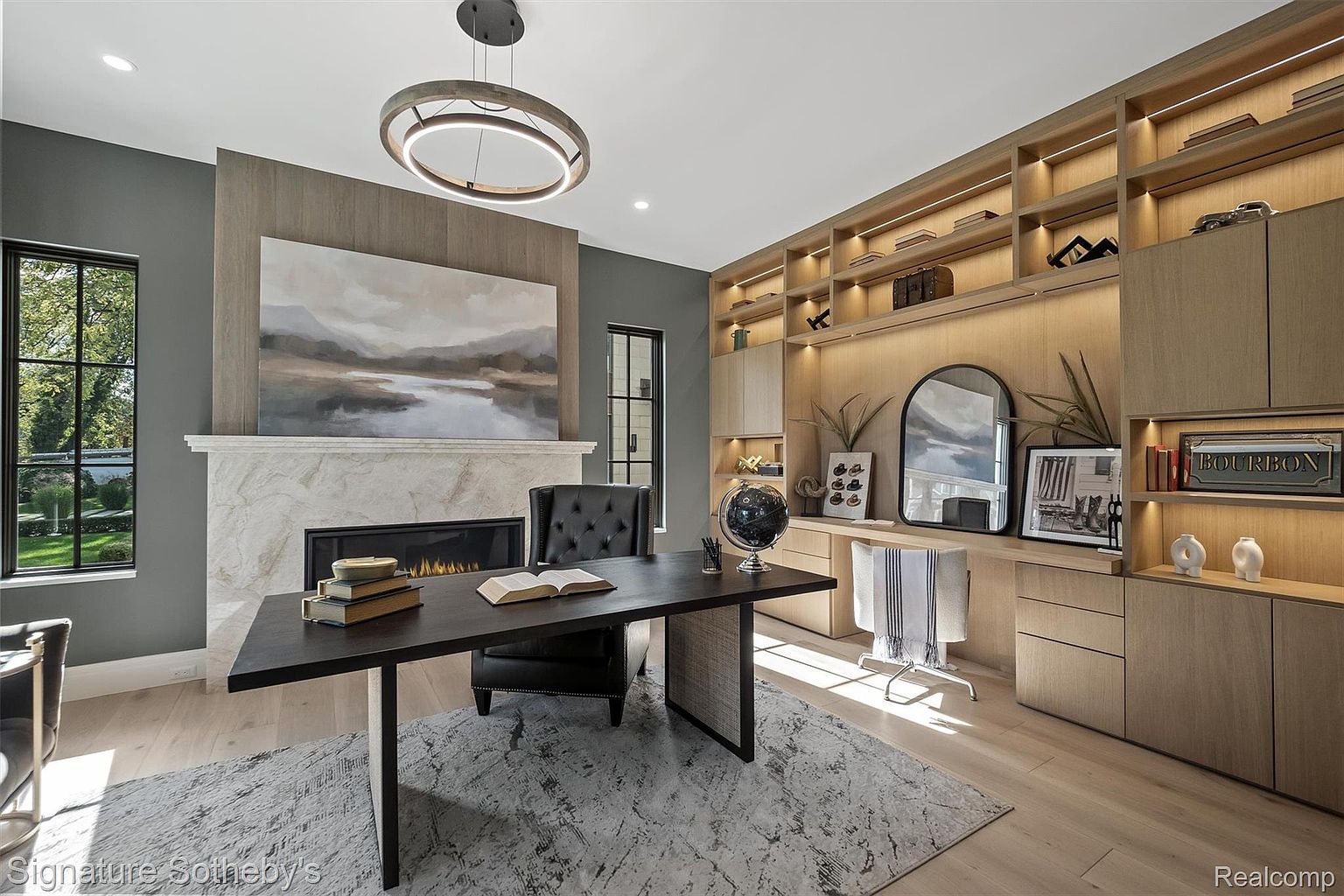 This is a well-appointed home office featuring a dark wood desk with books, a comfortable leather chair, and a fireplace with a marble surround. Custom built-in shelving provides ample storage and display space, illuminated by accent lighting. The room is decorated in neutral tones with a large abstract painting above the fireplace, creating a sophisticated and productive atmosphere.
