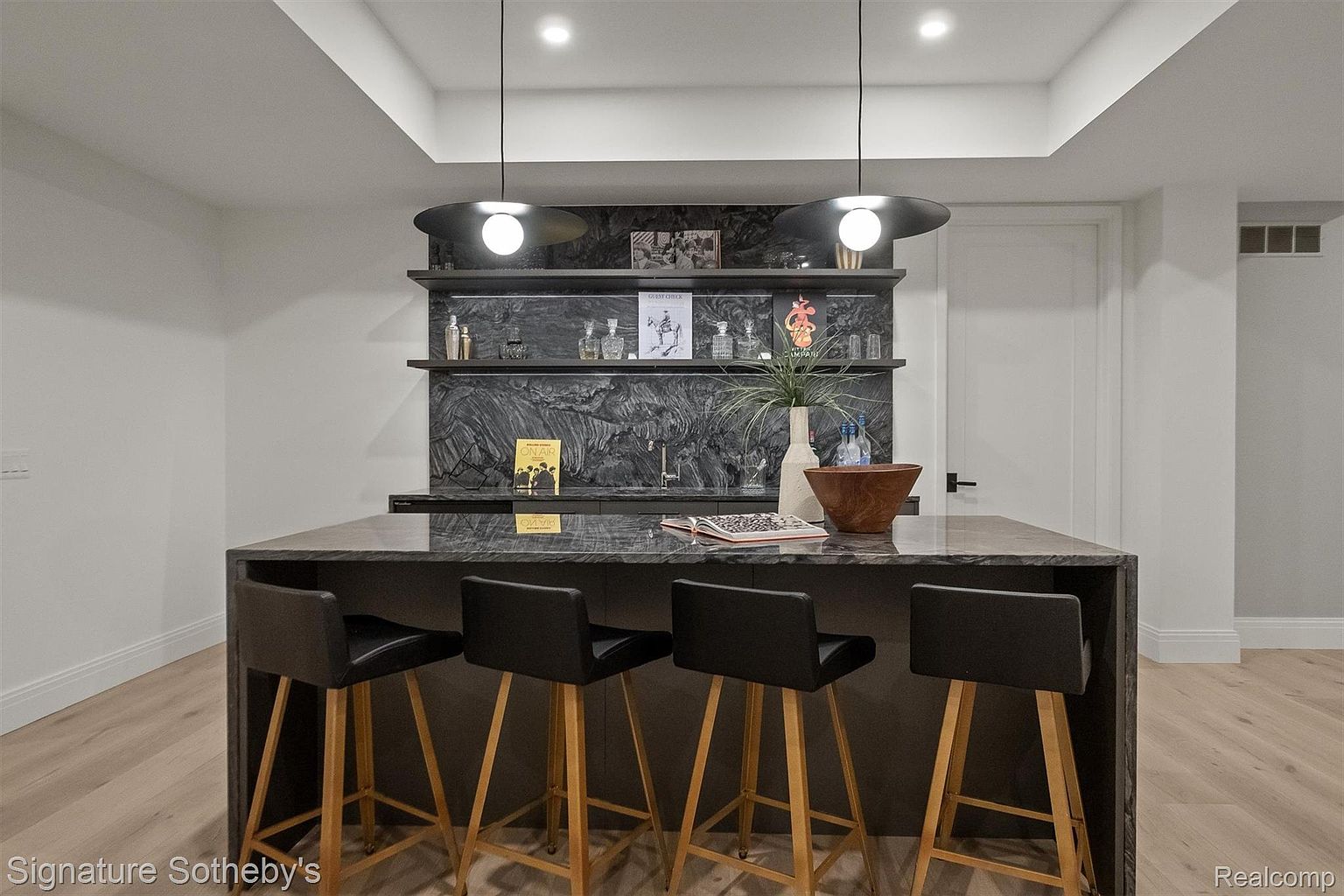 This interior shot showcases a stylish kitchen bar area with a dark marble countertop and backsplash, complemented by wooden-legged bar stools with black leather seats. Two modern pendant lights hang above the island, illuminating the space and highlighting the decorative items on the shelves behind the bar. The overall impression is modern and sophisticated.